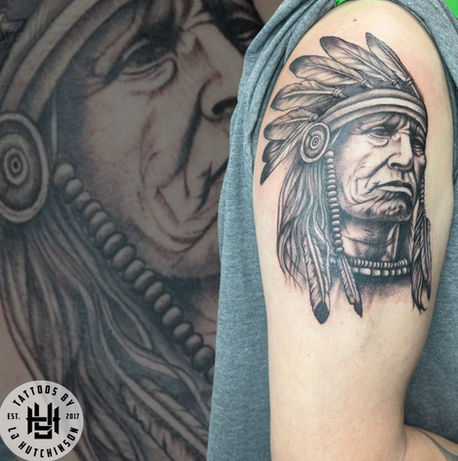Black and grey tattoo of an elder wearing a feathered headdress, done on the upper arm.