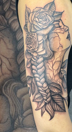 Tattoo of a woman’s cracked face with roses and braid, done in black and grey on the upper arm.