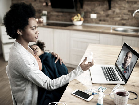 A woman sitting at a table, holding a baby in one arm while typing on a laptop with the other, representing quiet, multitasking leadership and the unseen work of mothers and caregivers.