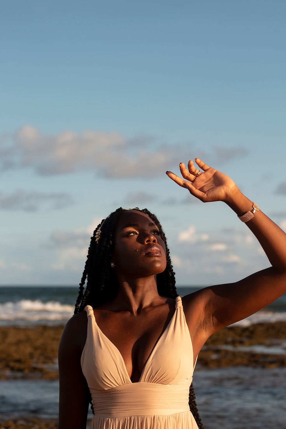 Black woman in a flowing white sundress shielding her eyes from the bright summer sun on a sandy beach, embodying peace, strength, and natural beauty.