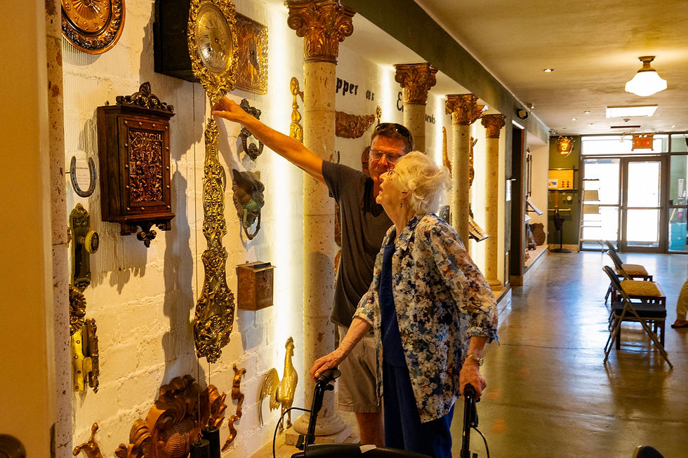 Man points at ornate wall decor in a warmly lit hallway. Elderly woman with walker observes, wearing floral jacket. Cozy, curious mood.