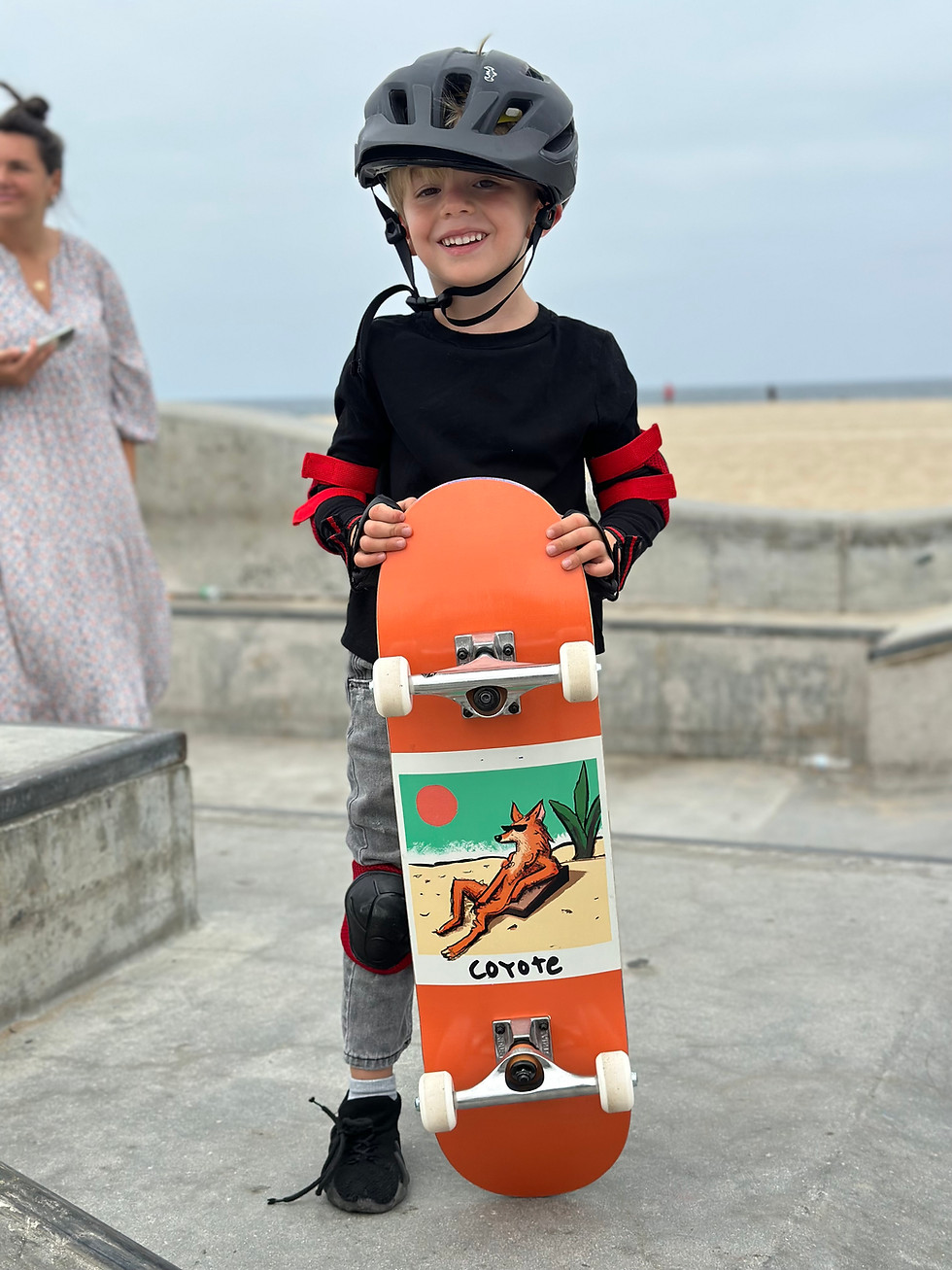 A kid smiling and holding his mini skateboard at the Venice Skatepark after his skateboarding lesson