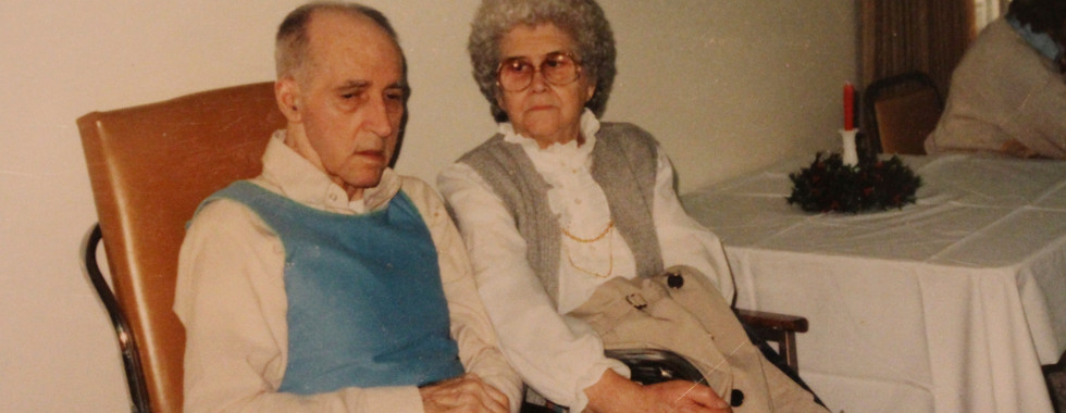 An elderly man in a wheelchair sits next to his wife in a nursing home lobby.