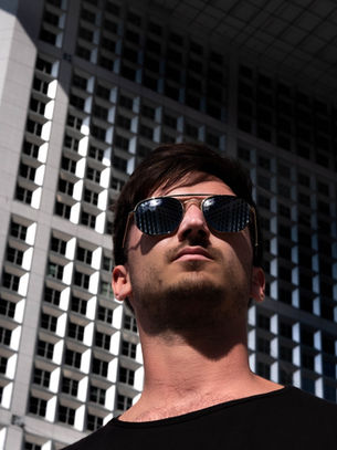 Man in sunglasses looking up at a grid-patterned building, reflecting in his lenses. He wears a black shirt; the mood is confident.