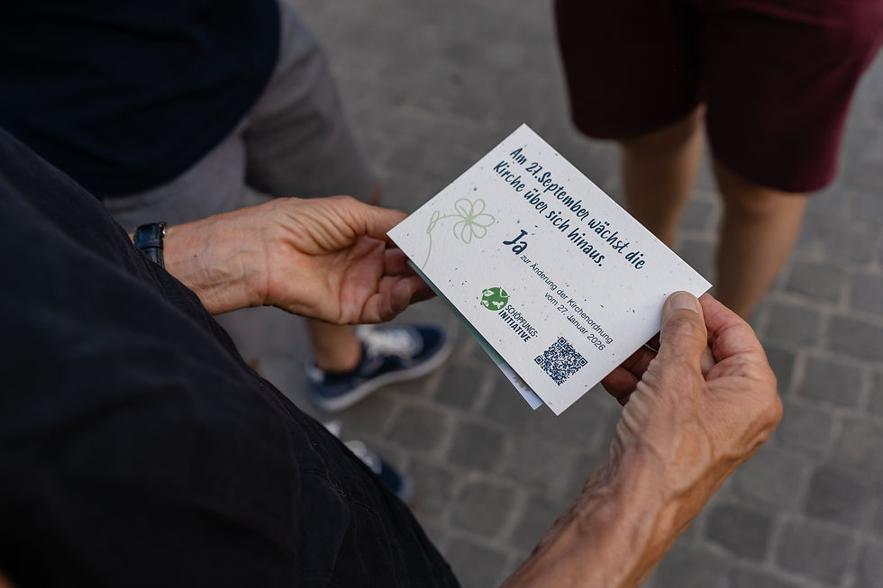 Ein Detailshot über die Schulter einer Person, die einen Flyer in der Hand hält. Darauf steht "Am 27. September wächst die Kirche über sich hinaus. Ja zur Änderung der Kirchenordnung vom 27. Januar 2026". Darunter ist eine Blume, das Logo der Schöpfungsinitiative und ein QR-Code.
