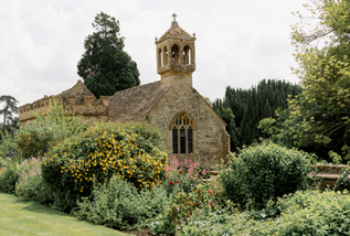 Small stone chapel with bell tower behind flowering garden.