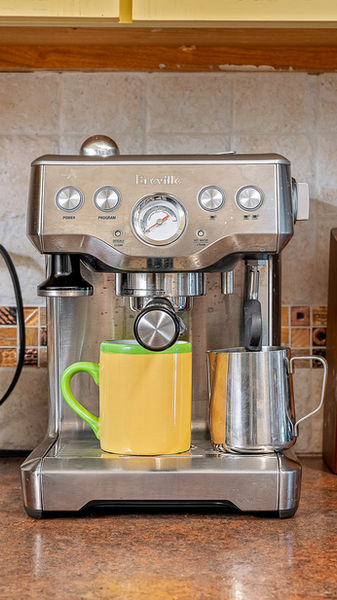 A breville espresso machine with yellow mug on the counter between a coffee can and  a knife set.