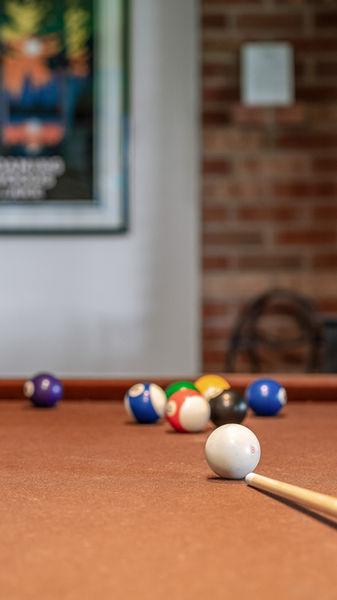 Brown felt pool table with balls and a cue. A woodstove and brick hearth in the backdrop with wall art nearby.