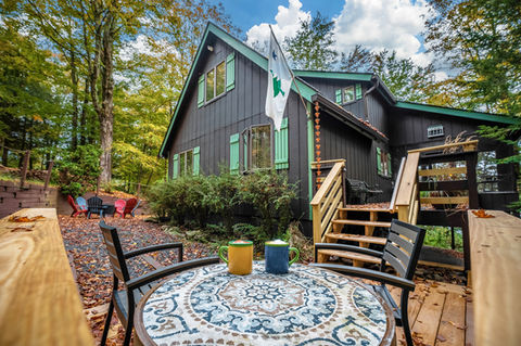 Tile table with coffee cups, two metal chairs, with brown cottage and green trip behind. 