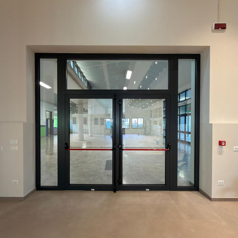 Vista interna di una porta a doppia anta in alluminio grigio antracite con maniglioni antipanico rossi, ampie vetrate laterali e sopraluce in un ambiente spazioso.