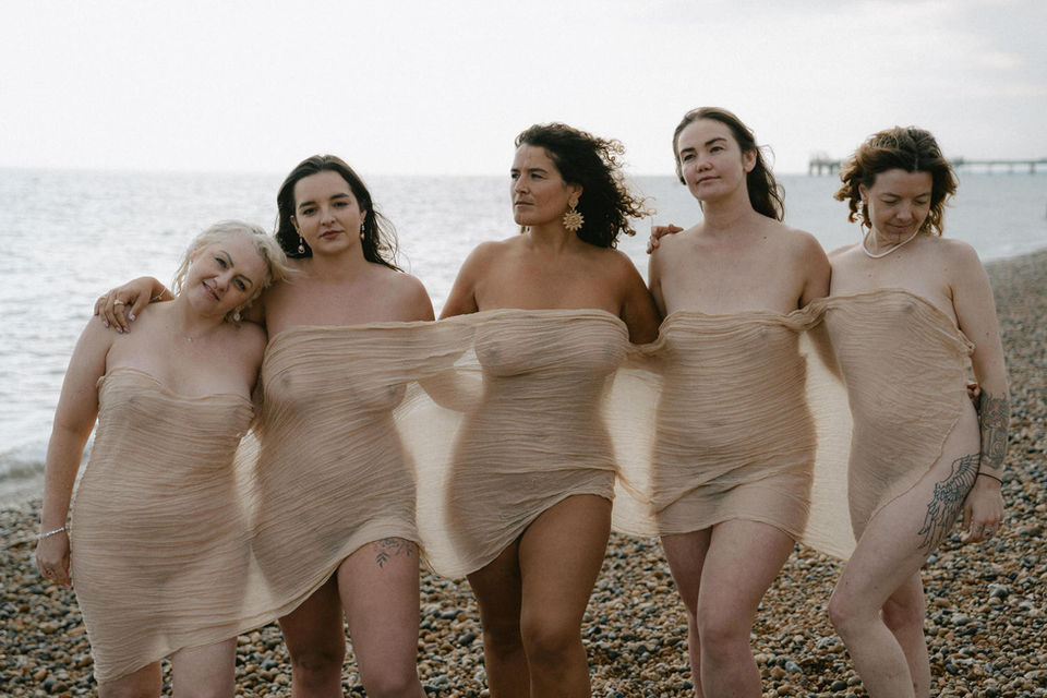 Five women of different body types wrapped in sheer fabric standing together on a pebble beach