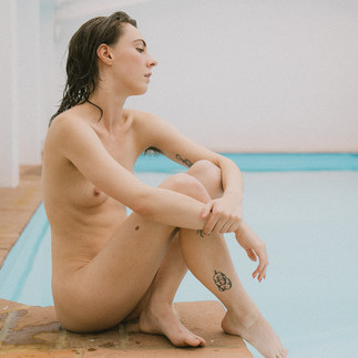 Poppy sitting naked on the side of the pool, looking away from the camera and hugging her knees