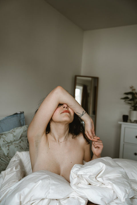 Woman sitting up in bed with arms raised above her head during an intimate portrait session