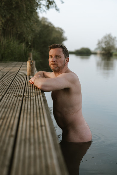 Naked man standing in a river leaning on a wooden jetty, looking over his shoulder