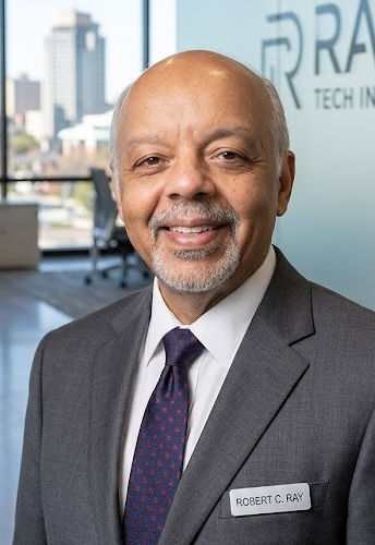 A man in a gray suit with a name tag smiles in an office setting. A cityscape view is visible through a window.
