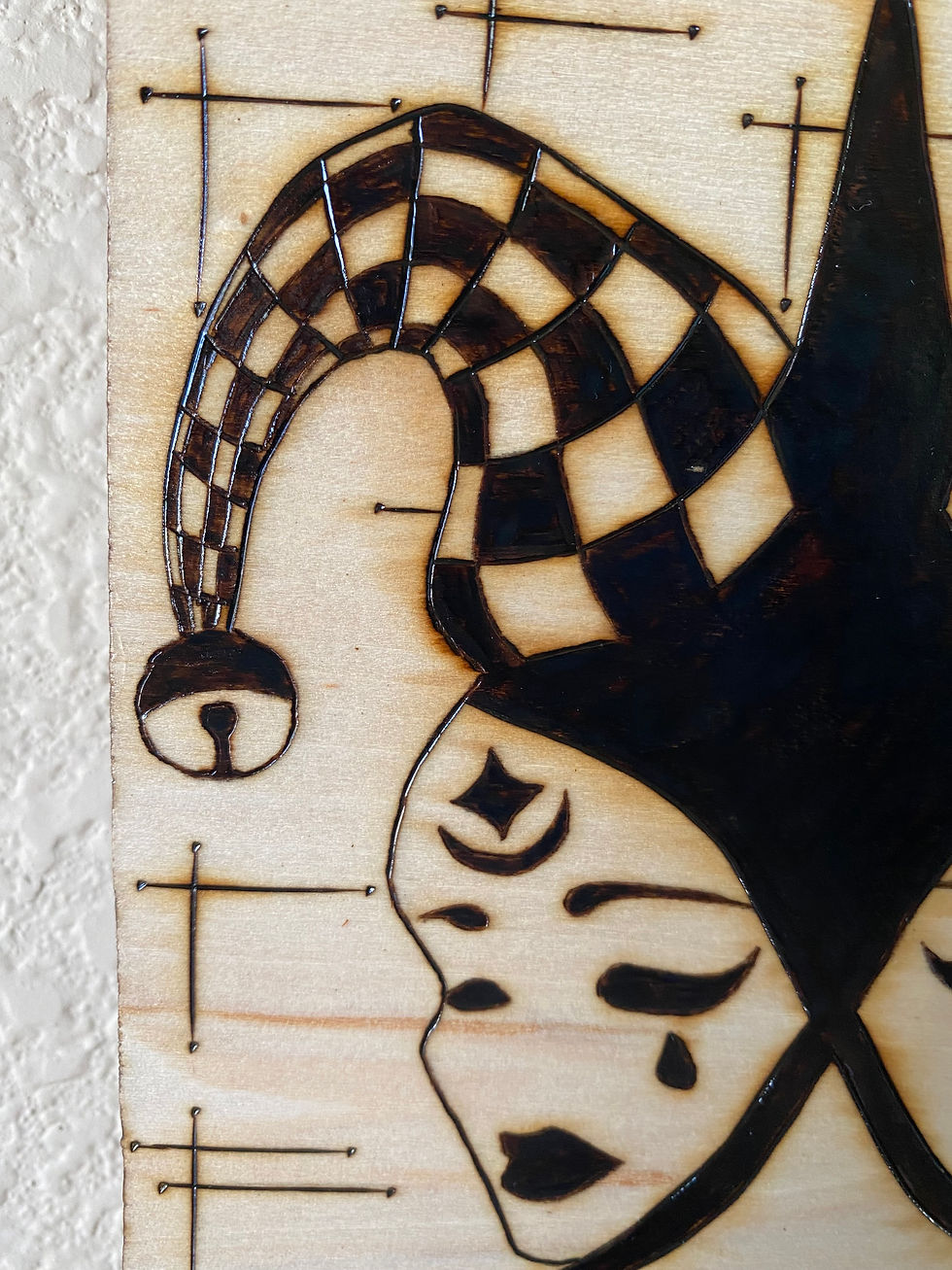 Thumbnail: Pyrography artwork: Jester mask with checkered hat and tearful faces.close up image of the left face