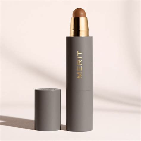 MERIT Beauty complexion stick with gold branding, taupe matte packaging, and cream tip for face makeup, shown on a neutral background with soft lighting.