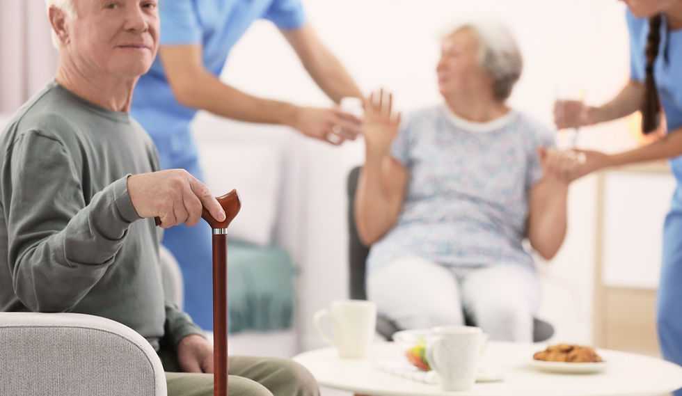 Elderly man with a cane sits in a care home while nurses and another senior woman chat nearby over tea and cookies