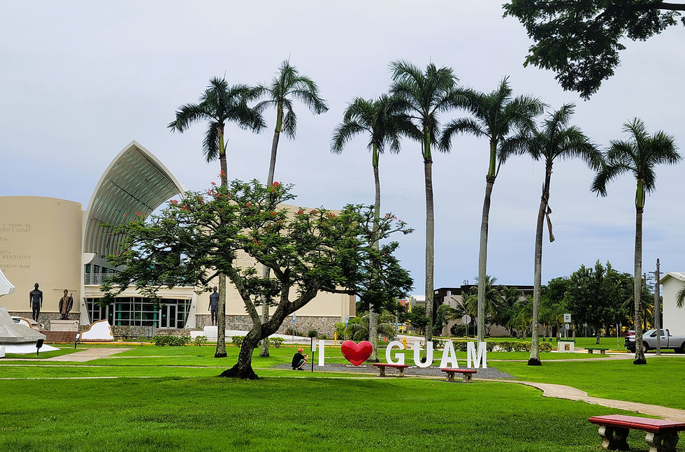 Lush park in Guam with palm trees, "I ❤️ GUAM" sign, and a modern building. A person sits on a bench, enjoying the peaceful setting.