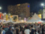 Crowded night street market with people under white tents, surrounded by lit palm trees and buildings. Vibrant and bustling atmosphere.