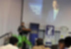 Man speaks at podium; screen shows "Honorable BJ Cruz - Public Auditor of Guam." Event banners and plants decorate the stage.