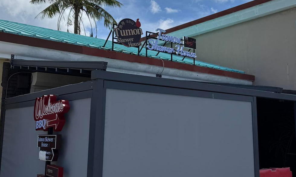 Sign for Tumon Skewer House with a red arrow and "Welcome BBQ", set against a turquoise roof and palm trees under a blue sky.