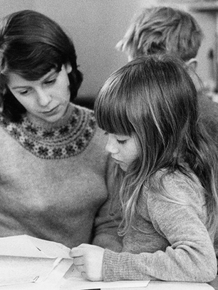 Black and white image of a woman sat looking at papers with a young girl on her lap and a boy stood behind her
