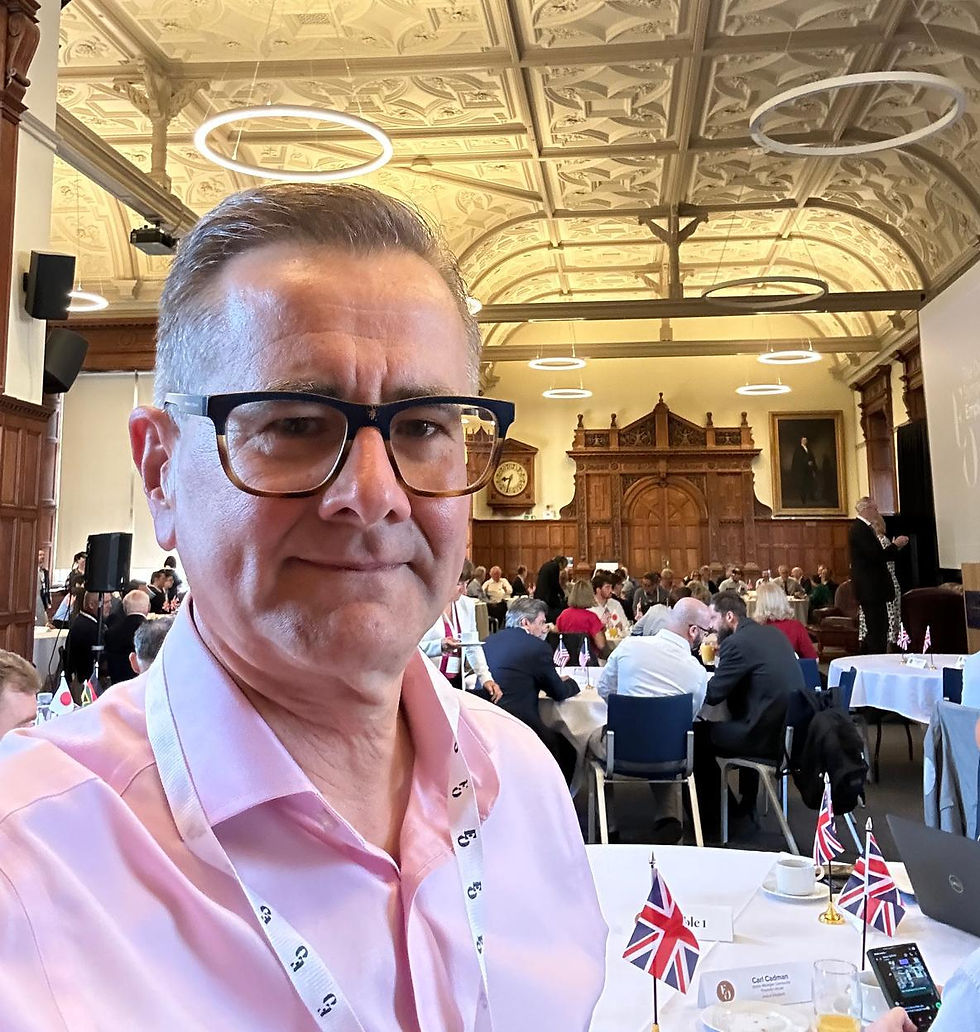Man in pink shirt and glasses at a formal event with ornate ceiling. Union flags and name card on table. Attendees in background.