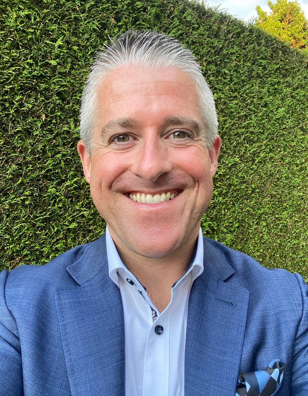 Smiling man in a blue suit with a patterned pocket square stands against a green hedge background.