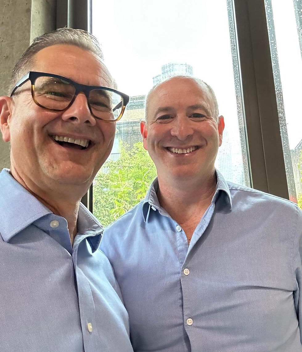 Two smiling men in blue shirts looking straight to camera, with a bright window showing a cityscape and greenery in the background.