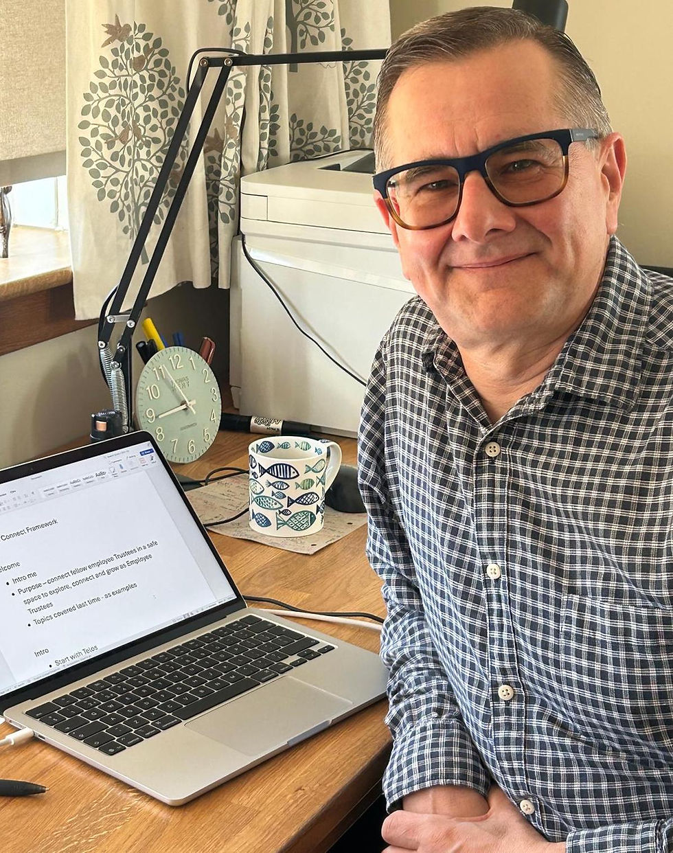 Man in glasses smiling at a desk with a laptop displaying text, a patterned mug and a clock. The room has leafy curtains.