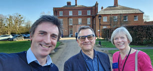 Three people smiling outdoors with a historic red-brick building in the background. Clear sky, greenery, and parked cars complete the scene.