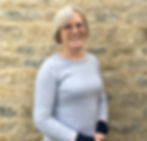Smiling woman in pale grey jumper looking at camera with sandstone wall behind.