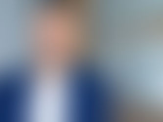 Smiling man in a blue suit and white shirt leaning against a glass office wall.