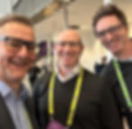 Three smiling men wearing glasses at an indoor event. They have speaker lanyards and are in a busy conference hall.