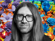 Artist Stuart Temple in black and white in front of a vibrant montage of his work "Bloom" currently at Jersey Zoon. You can see all the colours of the rainbow in the flowers which are biodegradable.