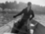 Jen Burden smiling on a boat while crabbing, holding a rope and wearing a cap and gloves. A forested shore is visible in the background, with calm water beneath an overcast sky in Washington or Oregon