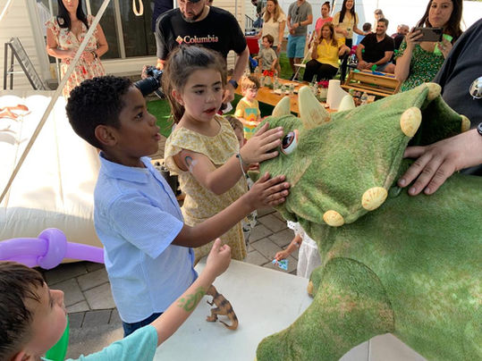 A dinosaur pet at a fair, a large, friendly dinosaur costume attracting the attention of children and families as they interact and take photos with it