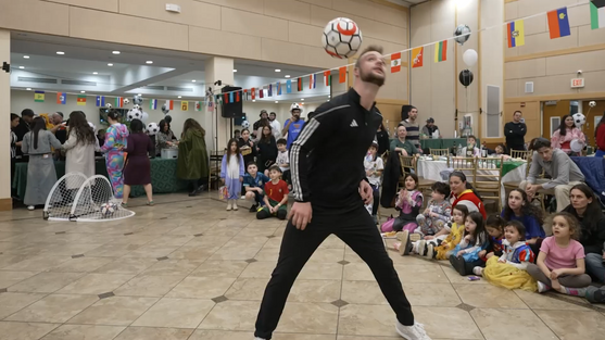 A professional soccer trick performer balancing a soccer ball on his head in front of attendees at a decorated fan festival.