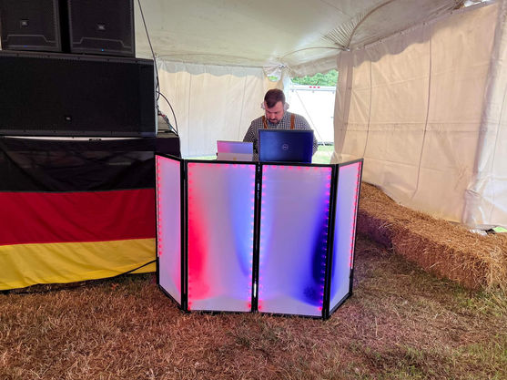 A professional DJ performing behind an illuminated red and blue LED facade booth with a German flag backdrop at an Oktoberfest event.