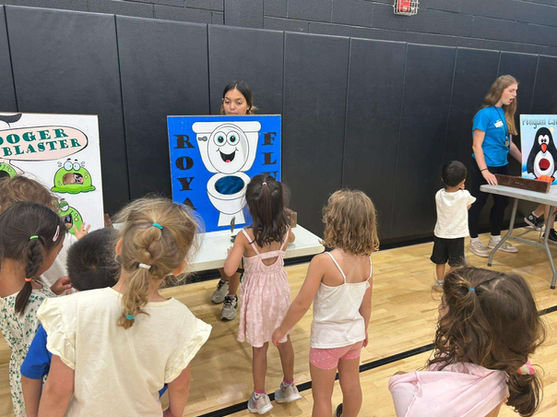 Children gathered around tabletop carnival game stations including Royal Flush and Booger Blaster during a Purim celebration.
