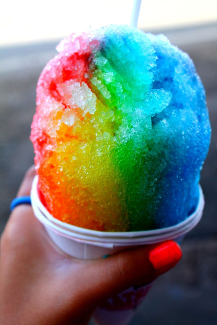 A close-up shot of a brightly colored rainbow snow cone served in a cup at an outdoor event refreshment booth.