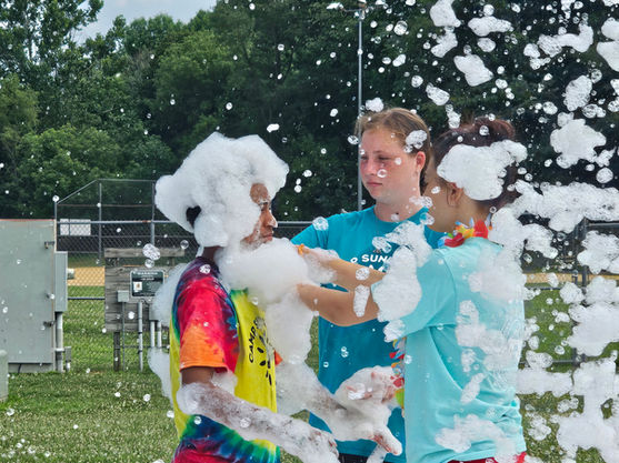 Two teenage girls in blue shirts placing thick foam suds onto a boy's head and neck during an outdoor youth foam event.
