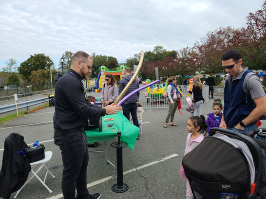 A man twists balloons into shapes while children and parents watch at an outdoor fair.