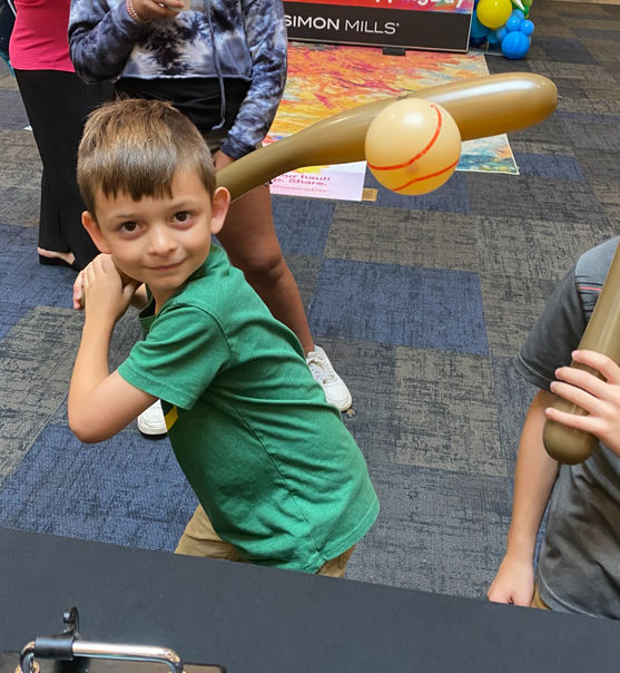 A young boy holding a brown balloon baseball bat and yellow balloon baseball made by a professional balloon twister at a sports fan festival.