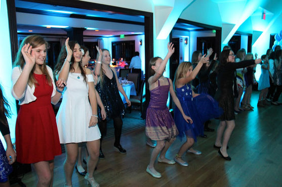 Young girls in colorful dresses dancing together under blue event lighting at a Bat Mitzvah DJ party.