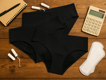 Beige calculator, tampons, pads, and three pairs of black affordable period underwear on a wooden table, showing cost comparison of menstrual products.
