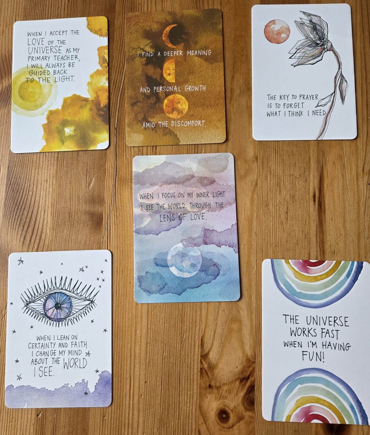 Zes spirituele kaarten met quotes en maan-, oog- en regenboogillustraties op een houten tafel.