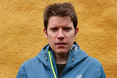 Musician James Brady looks directly into the camera with a neutral expression. He is wearing a blue-grey rain jacket and is standing against a textured yellow stone background.