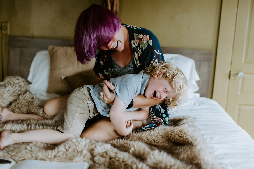 Mom and Son play on bed. Both laugh. Mom has purple hair covering her eyes and son has curly blonde hair. 